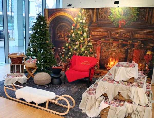 Textil-Fotodisplay mit Kaminzimmer-Motiv im Glasfoyer: Weihnachtsbaum, zwei Holzschlitten vorne, Sessel, Geschenkpakete rechts